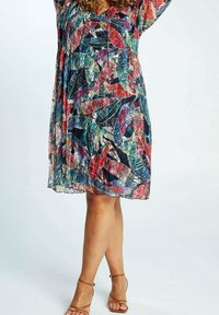 Nainen, joka on pukeutunut polvipituiseen, moniväriseen lehtikuosiseksi mekkoonsa, jossa on läpinäkyvää kangasta, ja vaaleisiin remmikenkiin korkokenkiä, seisoo jalat ristissä.