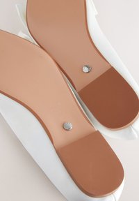 Dos zapatos planos blancos con suelas lisas de color beige y pequeños tachones metálicos circulares cerca de la zona del talón, colocados uno al lado del otro.