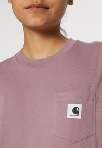 T-shirt a girocollo di colore viola chiaro con maniche corte e una tasca sul petto. La tasca presenta un patch del logo Carhartt in bianco e nero. Tessuto morbido.