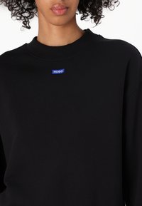 Schwarzes Sweatshirt aus weichem Material, mit einem gerippten Rundhalsausschnitt und einem blauen "HUGO"-Logo, das auf der Brust gestickt ist.