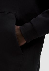 Dunkelhäutige Hand teilweise in der vorderen Tasche eines schwarzen Hoodies, wobei das gerippte Bündchen am Ärmel sichtbar ist.