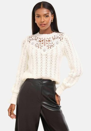 Weißer Strickpullover mit einer Spitzenblende, komplizierten Mustern und langen Ärmeln. Kombiniert mit hoch taillierten schwarzen Lederhosen.