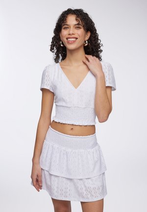 Completo bianco composto da due pezzi, caratterizzato da un top corto a maniche a sbuffo con scollatura a V e una gonna a balze smock. Tessuto in cotone con traforature e motivi delicati.