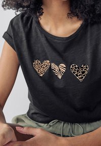 Sort t-shirt med tre hjerte design: et med leopardprint, et med zebra striber, og et med et mørkere leopardmønster.