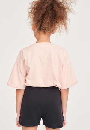 Kind mit lockigem Haar, das ein hellrosa übergroßes T-Shirt und schwarze Shorts trägt, steht mit dem Rücken zur Kamera vor einem einfarbigen hellen Hintergrund.