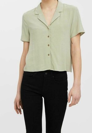 Persona con una camisa de manga corta color verde claro y pantalones negros de cintura alta sobre un fondo blanco.