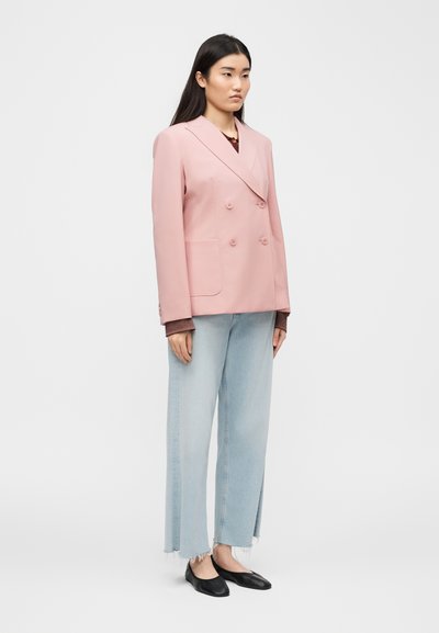 Γυναίκα με ροζ σακάκι διπλής σειράς κουμπιών, τζιν με ξεφτισμένο τελείωμα σε ανοιχτό μπλε, καφέ μπλούζα με μακριά μανίκια και μαύρες μπαλαρίνες, στέκεται σε ουδέτερο χώρο.
