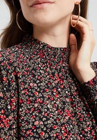 Svart blommig topp med samlad halsringning och långa ärmar; röda och beige blommor mot en mörk bakgrund. Guldörhängen i form av ringar syns.