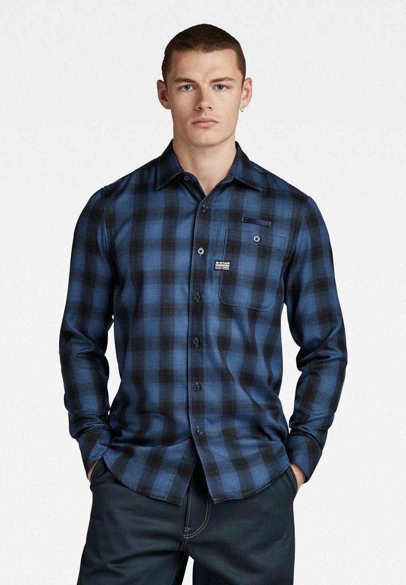 Blauwe en zwarte geruite blouse met lange mouwen, knopen aan de voorkant, één borstzak en een klein merklabel met een zachte textuur.