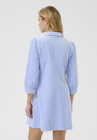 Una donna con lunghi capelli biondi che indossa un vestito azzurro chiaro a costine con maniche a tre quarti, vista di spalle su uno sfondo neutro.