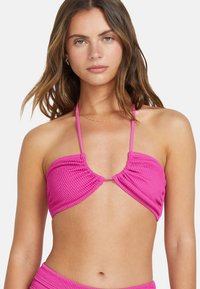 Ružový pletený bikiny top s viazaným halter dizajnom, ktorý má zhrnutú textúru a výrez v strede. Bez ďalších akcentov.