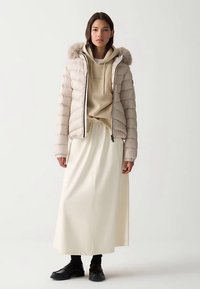 Giacca imbottita beige con cappuccio foderato di pelliccia, con chiusura a zip e polsini a costine, indossata sopra una felpa con cappuccio beige e una gonna bianca.