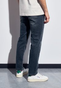 Persona de pie en interiores, vistiendo jeans azul oscuro, zapatillas blancas con tacones verdes y una camisa de color claro.