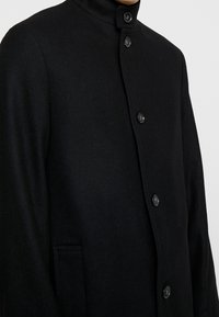 Mustat villakangastakki, jossa on korkea kaulus, nappikiinnitys edessä ja sivutaskut. Kankaan pinta on sileä ja muotoiltu.