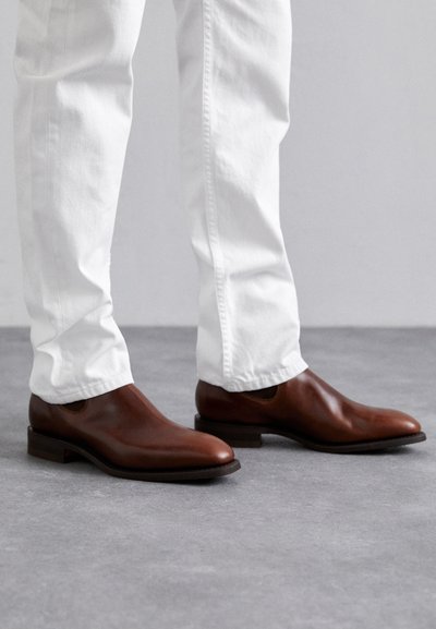 Brown leather slip-on shoes with a smooth finish, pointed toe, and flat sole, paired with white slim-fit pants.