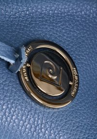 Dettaglio di una borsa in pelle blu con un logo rotondo in tono oro con un design a spirale e superficie testurizzata, accentuata da una tracolla di un blu chiaro.