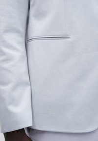 Blazer bleu clair avec un design élégant, un tissu lisse et une poche cachée avec un détail de bordure subtile. Coupe ajustée et structurée.