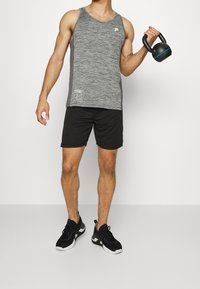 Canotta sportiva grigia con pannelli laterali grigi scuri, abbinata a pantaloni corti neri. La persona tiene in mano un kettlebell nero con accentuazioni blu. Sneakers nere.