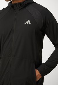 Zwarte sportjack met capuchon, heeft een grijs logo, rits aan de voorkant, elastische manchetten en gestructureerde panelen voor verbeterde ademend vermogen.