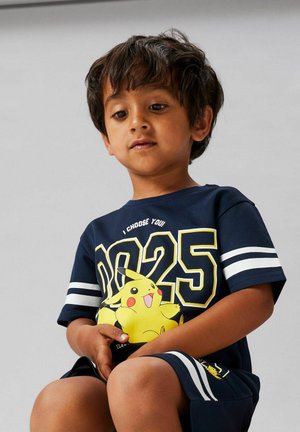 Jeune garçon portant une tenue bleu marine à thème Pikachu avec le numéro 025 et la phrase « Je te choisis ! », assis et regardant vers le bas.