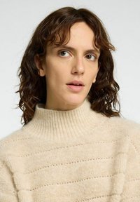 Stickad tröja i beige med en hög ribbad krage, med en mjuk textur och ett horisontellt randigt mönster med små perforeringar.