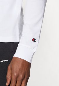 Camicia bianca a maniche lunghe con polsino a coste, caratterizzata da un piccolo logo Champion rosso e nero sul polso. Materiale in cotone liscio.