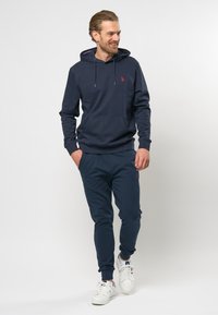 Marineblå bomullshoodie med frontlomme, sølvfargede snorer og liten rød logo. Kombinert med matchende joggebukser og hvite joggesko.