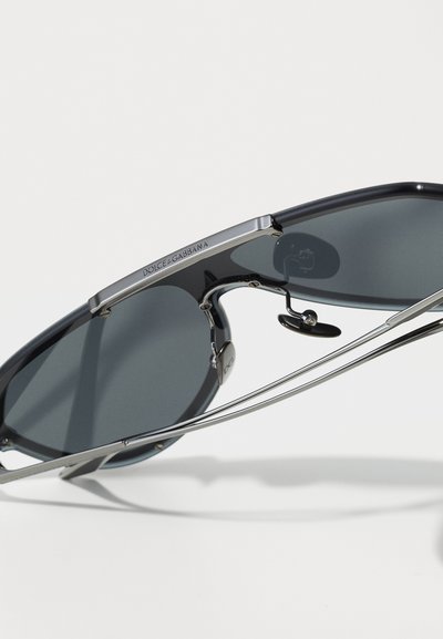 Dolce & Gabbana saules brilles ar tumši pelēkiem stikliem, gludu melnu plastmasas rāmi un sudraba metāla akcentiem; iekļauj tiltu un deguna spilventiņus.