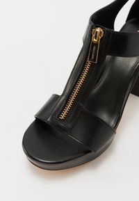 Sandale en cuir noir avec une fermeture éclair dorée à l'avant et un design à bout ouvert, avec un talon épais et une large bride à l'avant.