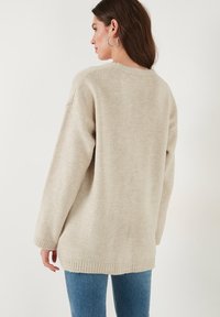 Maglione lavorato a maglia beige con vestibilità ampia, scollo rotondo e polsini a coste. Texture morbida; arriva fino a metà coscia, senza motivi o disegni visibili.