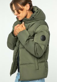 Oliwkowa kurtka puchowa z odpinanym kapturem, zamkiem z przodu i czarną łatą z logo na lewym rękawie; posiada duże pikowane panele.