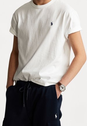 Witte katoenen t-shirt met opgerolde mouwen en een klein marineblauw logo op de borst, gecombineerd met marineblauwe broek en een zilveren horloge met een bruin leren band.