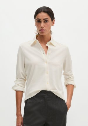 Vrouw draagt een crème lange mouwen blouse met opgerolde mouwen en donkergrijze hoge taille broek, staand met één hand in de zak.