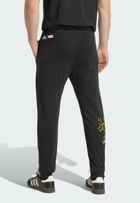 Pantalones deportivos negros de corte ajustado, con una cintura elástica y un diseño gráfico amarillo en la parte inferior de la pierna izquierda, combinados con zapatillas deportivas negras.