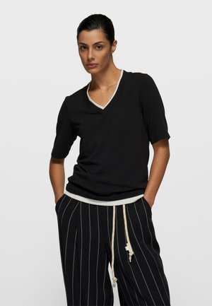 Zwart T-shirt met korte mouwen en V-hals, voorzien van een witte bies en een subtiel geborduurd ontwerp op de borst. Gecombineerd met gestreepte zwarte broek.