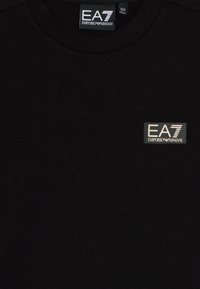 Fekete pamut póló, kerek nyakkivágással és téglalap alakú logócímkével, ami az "EA7" és az "Emporio Armani" feliratokat ezüst és fehér betűkkel tartalmazza.