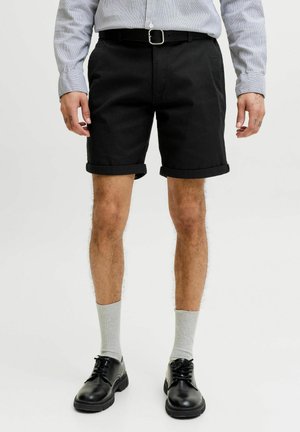 Muž nosiaci čierne šortky s vyhrnutými lemami, pruhované tričko s dlhým rukávom zastrčené dnu, sivé ponožky, čierne kožené topánky a čierny opasok.