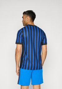 Chemise à manches courtes noire et bleue à rayures verticales avec un tissu texturé. Associée à un short bleu, mettant en valeur un design sportif.