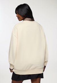 Il pullover oversized crema presenta un tessuto morbido, spalle cadenti, polsini a coste e un colletto scuro a contrasto. Abbinato a una gonna nera.