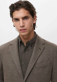 Blazer marrone a motivo con una trama strutturata, reverse e taschino sul petto, indossato sopra una camicia scura con bottoni e sottili righe verticali.
