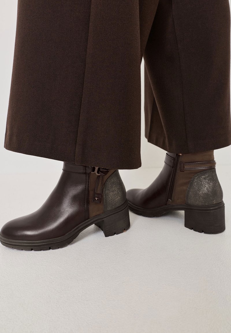 Gros plan sur une personne portant des bottines marron foncé à fermeture éclair latérale et détail de talon métallique, associées à un pantalon marron à jambes larges.