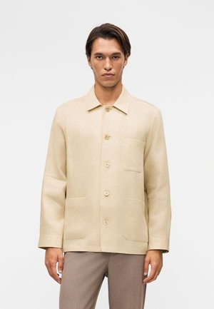 Vyras, vilkintis smėlio spalvos striukę su sagomis ir trimis priekinėmis kišenėmis, dėvi šviesiai rudus kelnes, stovintis priešais paprastą baltą foną.