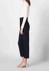 En kvinna står på sidan, iklädd en figurnära krämfärgad topp, högmidjade marinblå byxor och silverfärgade klacksandaler, vilket visar upp en modern outfit.