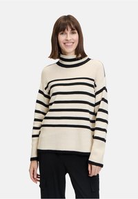 Cremfarbener Rollkragenpullover mit schwarzen horizontalen Streifen, gerippter Textur, langen Ärmeln und lässiger Passform, kombiniert mit dunklen Hosen.