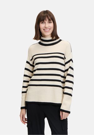 Pull col roulé crème avec des rayures horizontales noires, texture côtelée, manches longues et coupe décontractée, associé à un pantalon sombre.