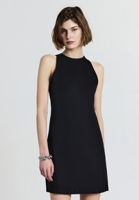 Abito nero senza maniche con un collo alto, tessuto liscio, silhouette a A e un leggero lucido. Accentuato da un bracciale d'argento al polso.