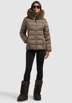 Femme portant une doudoune marron avec un grand col en fourrure, des leggings noirs, des bottes d’hiver noires et des lunettes de soleil foncées, debout sur un fond uni.