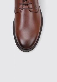 Zapato de vestir de cuero marrón con una textura suave, punta redonda y suela de goma negra. Presenta costuras tonales y cierre con cordones.