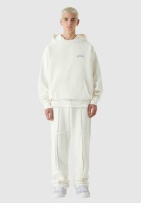Sudadera blanca con bolsillo frontal, presenta un sutil logo morado. Combinada con pantalones de forro polar blancos a juego y zapatillas blancas.