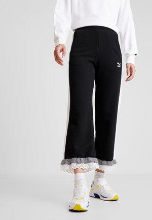 Persoon die zwarte cropped broek draagt met witte zijstrepen en kantafwerking, witte sokken, witte sneakers met gele accenten en een wit sweatshirt.